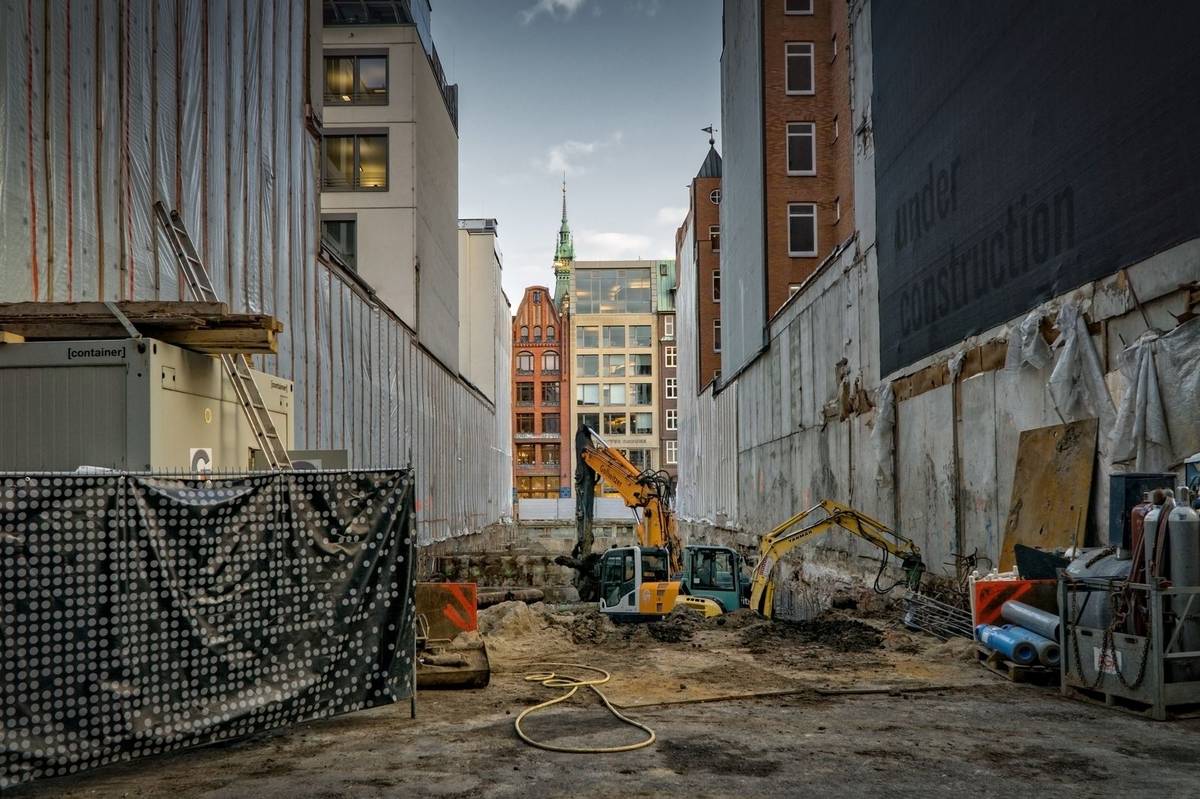 Two tracked excavators working a basement excavation between sheet-piled retaining walls on an inner-city site, with spoil heaps, a container and a hose across the mud