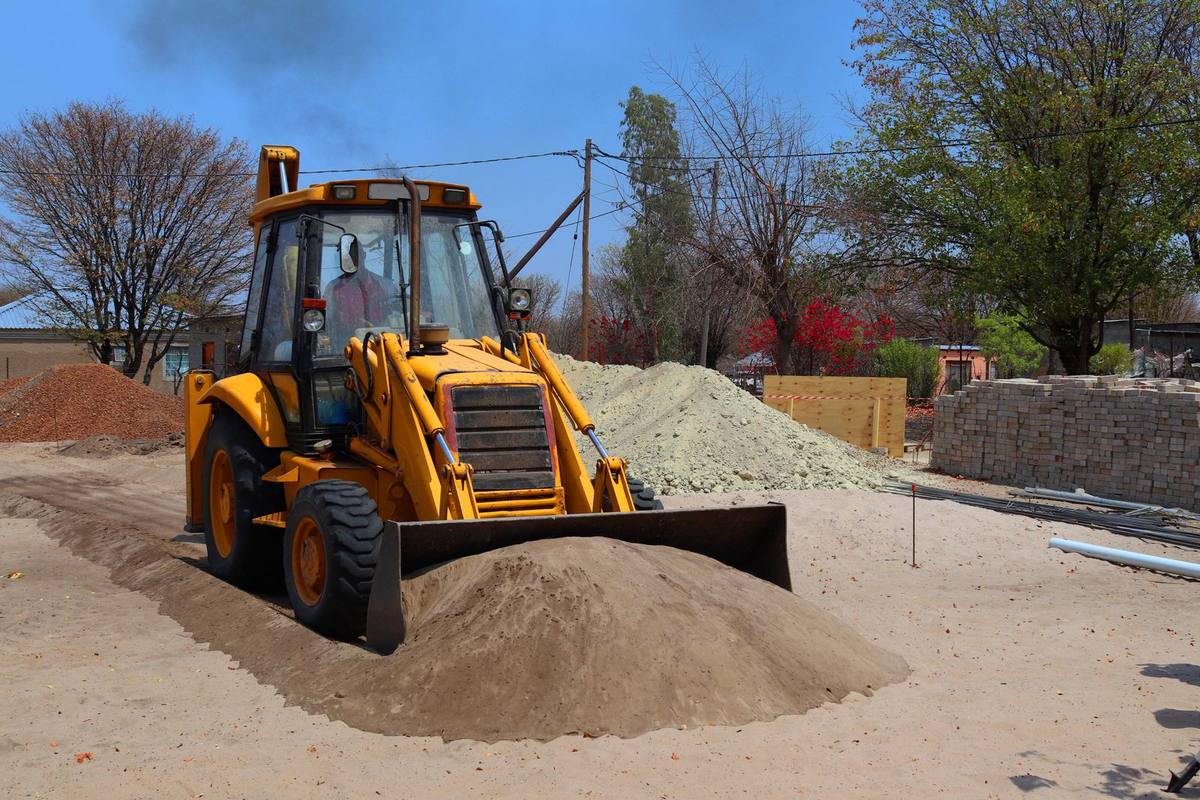 A yellow backhoe loader levelling a sand heap on a house plot, with a part-built brick wall, sand and stone heaps and a boundary wall behind it